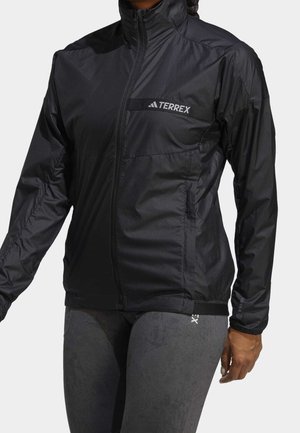 Veste Terrex noire zippée avec col montant portée sur un legging gris, visible de la poitrine jusqu'à la moitié de la cuisse sur une personne debout.