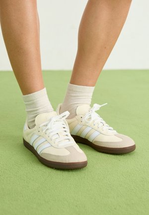 Para nóg w beżowych butach Adidas Samba z białymi paskami i jasnobeżowych skarpetkach stojąca na zielonym dywanie.