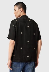 Mann som har på seg en svart kortermet skjorte med små hvite prestekrager og blå jeans, vendt bort fra kameraet mot en ensfarget bakgrunn.