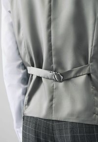 Gilet satin gris avec un design ajusté, présentant une boucle dans le dos et une texture lisse, associé à une chemise blanche et un pantalon à motifs.