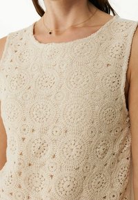 Haut beige en crochet présentant un motif floral circulaire ; finition texturée avec des mailles ouvertes, encolure arrondie et design sans manches.