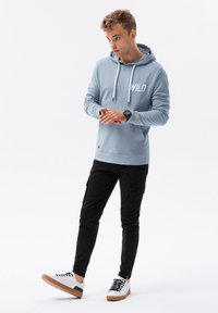 Felpa azzurra chiara con la scritta "WILD" stampata in bianco, abbinata a pantaloni cargo neri e sneakers bianche con suole marroni.