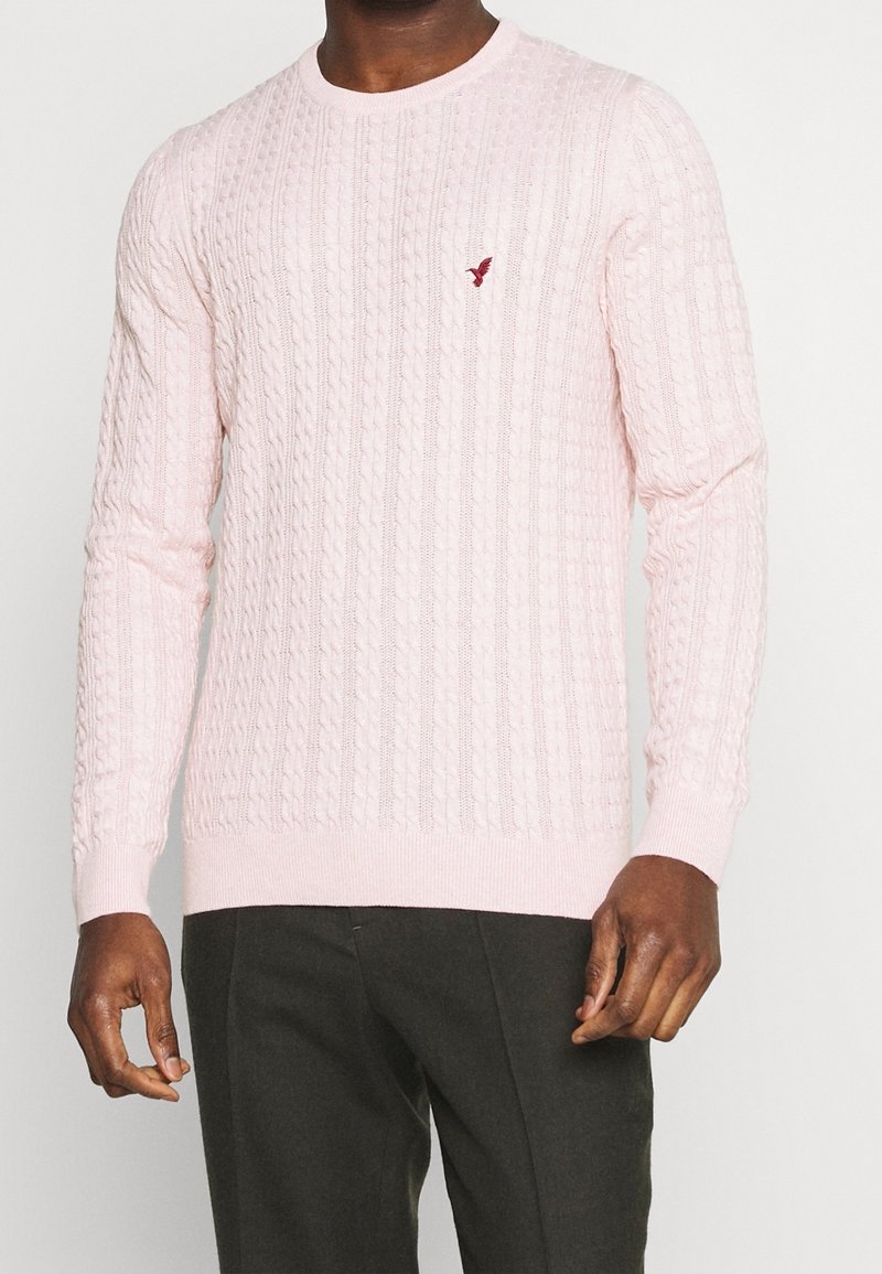 Light pink cable-knit sweater with a round neckline and long sleeves. Features a small, contrasting logo on the chest and ribbed cuffs.