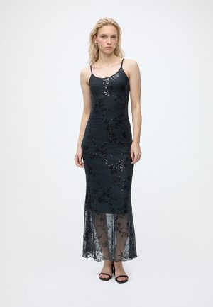 Femme portant une robe noire sans manches avec de la dentelle florale et des détails en sequins, debout devant un fond blanc avec des sandales à talons noires.