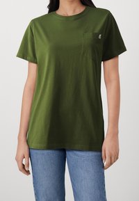 Camiseta de algodón color verde oliva con cuello redondo, mangas cortas y un bolsillo en el pecho. Presenta un pequeño logo en el bolsillo.
