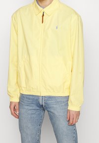 Hellgelbe Zip-Jacke aus Baumwolle mit spitzem Kragen, elastischen Bündchen und kleinem Logo auf der Brust.