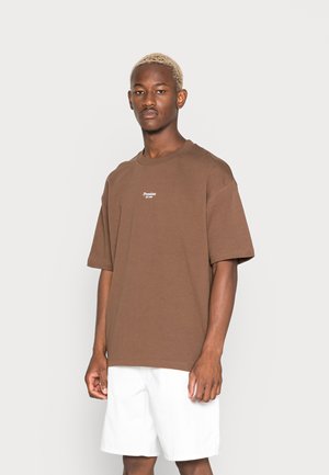 Brun bomull t-skjorte med rund hals og kortermet. Har en liten hvit logo på brystet. Bært med hvite shorts.