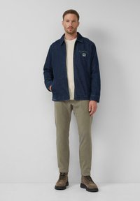 Mann, der gegen eine graue Wand steht und eine marineblaue Jacke, einen cremefarbenen Pullover, olivgrüne Hose und braune Schnürstiefel trägt, mit den Händen in den Jackentaschen.
