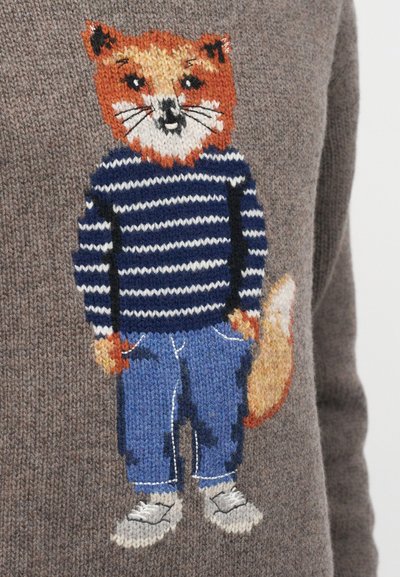 Adīts lapsas attēls, kas ģērbta zilgani baltā svītrainā džemperī, zilās biksēs ar jostām un baltajās sporta apavos pelēkā auduma fona.