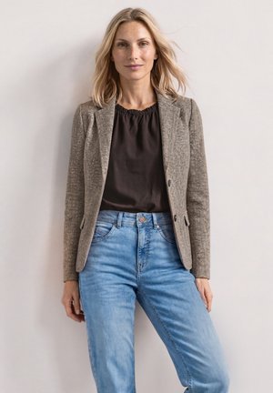 Blondynka ubrana w brązową teksturowaną marynarkę, czarną bluzkę i jasnoniebieskie dżinsy, stojąca na tle jednolitego jasnego tła.
