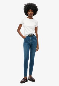 Jeans ajustados azul de cintura alta, hechos de mezclilla. Combinados con una camiseta blanca de manga corta y mocasines marrón oscuro. Detalle de cinturón simple.