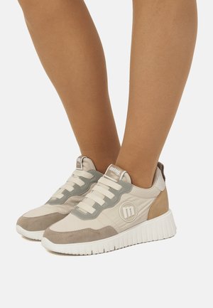 Zapatillas casuales beige y gris con suelas blancas, usadas en los pies sobre un fondo liso.