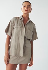 Khaki šatové šaty s krátkými rukávy, zapínání na knoflíky vpředu, dvě náprsní kapsy a přiléhavá sukně. Hladká tkanina, minimalistický design, jemný lesk.