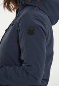 Laivansiniset ulkoilutakit, joissa on teksturoitu kangas, huppu ja pyöreä musta logopatchi ylälaidassa.