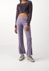 Person som bär lila sammetsbyxor med vida ben, grå sneakers och en långärmad genomskinlig kort topp med lila och svarta mönster, står på vit bakgrund.