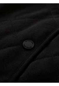 Zwart gewatteerde stof met een textuur. Heeft een zwarte knoop met het merk "TOM TAILOR". Genaaide lijnen vormen een ruitpatroon.