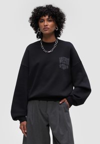 Felpa nera con un fit rilassato, caratterizzata da una grafica sul petto. Abbinata a pantaloni grigi a pieghe. Tessuto morbido al tatto.