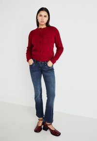 Cárdigan rojo de felpa con botones negros, combinado con jeans azules. La modelo lleva zapatos de tacón rojos brillantes, de pie frente a un fondo blanco.