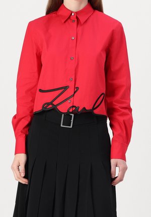 Vrouw die een rood overhemd draagt met zwarte "Karl"-tekst aan de zoom, gecombineerd met een zwarte geplooide rok en een riem met een vierkante gesp.