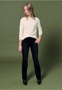 Eine Frau, die ein weißes Langarmshirt, schwarze Jeans und schwarze Stiefel trägt, steht vor einem grünen Hintergrund und schaut nach unten zur Seite.