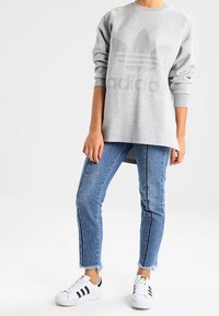 Felpa grigia con un grande logo tono su tono, abbinata a jeans in denim blu con strisce laterali e sneakers bianche con accenti neri.
