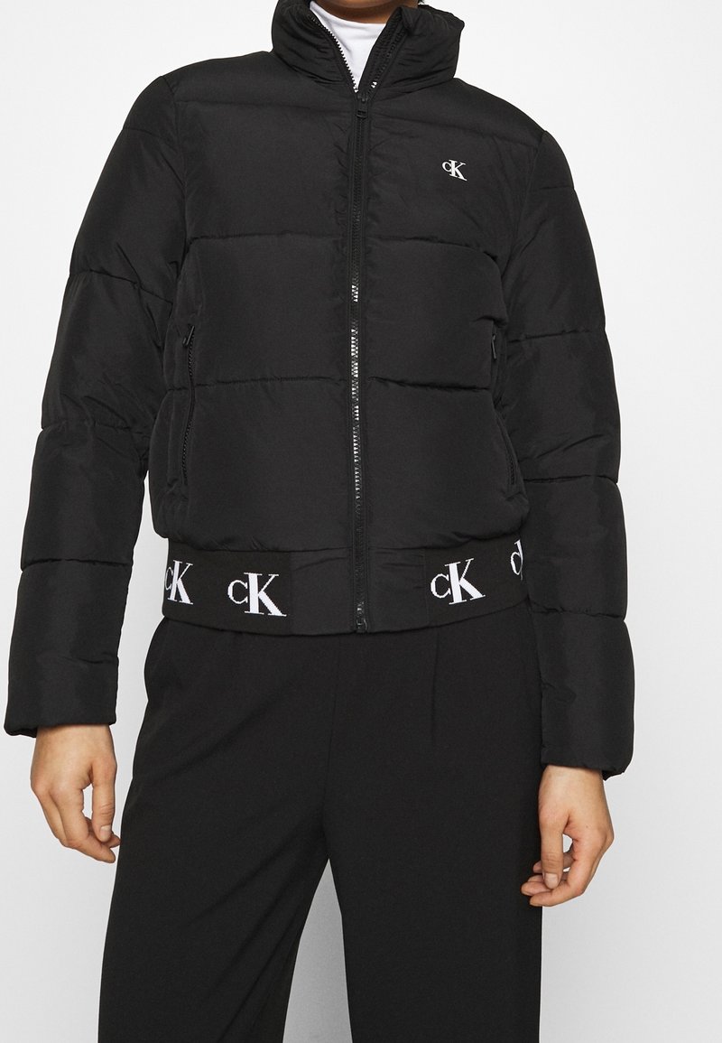 Black puffer jacket with a high collar, horizontal quilted design, zipper front, and elastic waistband featuring a white logo.