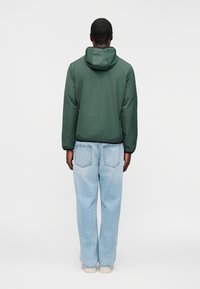 Άνδρας που στέκεται με την πλάτη γυρισμένη, φοράει σκούρο πράσινο μπουφάν με κουκούλα και ανοιχτό μπλε τζιν παντελόνι κανονικής γραμμής με λευκά παπούτσια.