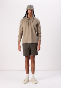 Polo beige à manches longues, shorts verts foncés, baskets beiges, et casquette gris clair ; tenue décontractée, texture lisse, design simple.