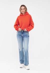 Rode oversized hoodie met een trekkoord voor de capuchon, gecombineerd met lichtblauwe flared jeans en witte sneakers op een effen witte achtergrond.