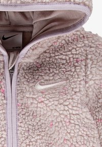 Fleece zip-up jakna v svetle bež barvi z rožnatimi pikicami, z kapuco in srebrnim Nike logotipom spredaj. Mehko na otip z rebrastimi robovi.
