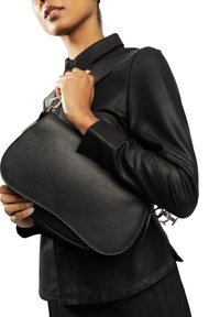 Borsa a mano in pelle nera con finitura testurizzata, forma curva e dettagli in metallo color oro, tenuta da una mano contro un abbigliamento nero.