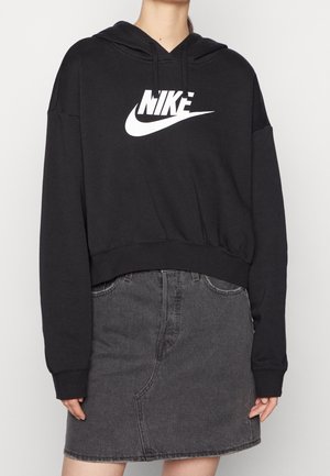 Persoon draagt een zwarte Nike-hoodie met wit logo en een donkergrijze denimrok, staand tegen een effen achtergrond.