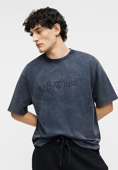 Νεαρός άνδρας με σγουρά μαλλιά κοιτάζει αριστερά, φορώντας μια ξεθωριασμένη σκούρα μπλε μπλούζα Off-White™ και μαύρο παντελόνι με κορδόνι, σε απλό φόντο.