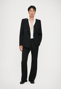 Jachetă de costum neagră cu două rânduri de nasturi, cu rever ascuțit, purtată cu o cămașă albă. Pantaloni negri cu croială lejeră și pantofi negri. Textură a materialului neted.