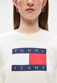 Κρεμ φούτερ με ορθογώνιο λογότυπο σε ναυτικό μπλε, λευκό και κόκκινο, που εμφανίζει το "TOMMY JEANS" με έντονα γράμματα.