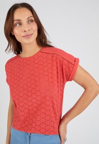 Top corto de manga corta rojo con textura perforada y un sutil patrón de puntos. Presenta un escote redondo y mangas enrolladas. Combinado con jeans de color azul claro.
