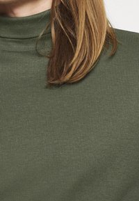 Kvinde med skulderlangt lyst brunt hår iført en ribbet olivengrøn turtleneck sweater, der er cropped ved halsen og skuldrene.