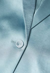 Blazer en velours bleu clair avec un col cranté, une fermeture à bouton rond et une texture lisse, mettant en valeur son design fluide.