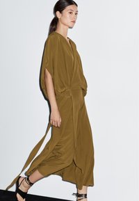 Olivově zelené zavinovací šaty s širokými rukávy, páskem s kulatou sponou a splývavým materiálem. Modelka je zobrazena z profilu.
