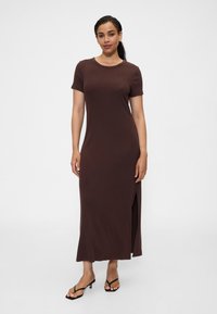 Brązowa, długa sukienka na krótkie rękawy z okrągłym dekoltem i bocznym rozcięciem, wykonana z miękkiego materiału. Modelka nosi czarne sandały na obcasie.