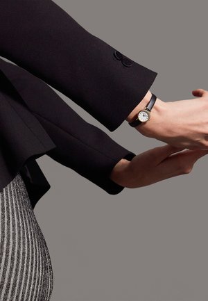 Sort blazerærme med knapdetalje, sølv- og sortstribet stof, guld-accentueret armbåndsur med hvid urskive og sort læderrem.