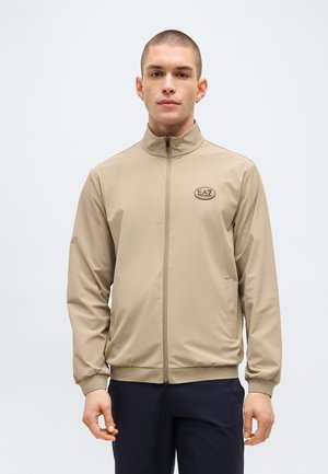 Rövid hajú férfi bézs színű, cipzáras EA7 logós dzsekit visel a bal mellkasán, tengerészkék nadrágban áll, világos egyszínű háttér előtt.