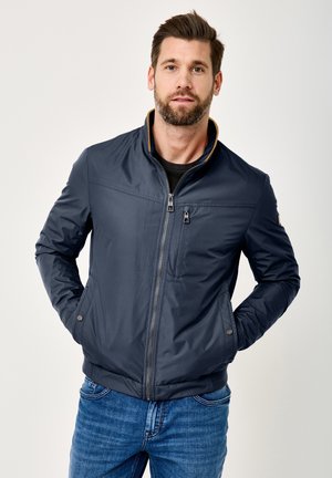 Man with beard wearing navy zip-up jacket and blue jeans, standing with hands in pockets against plain background.