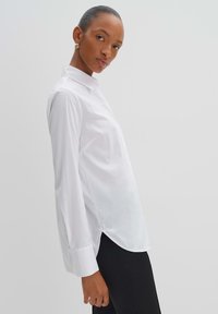 Camicia bianca a maniche lunghe con colletto a punta, caratterizzata da una vestibilità rilassata, fessure laterali e maniche con polsini, realizzata in tessuto leggero.