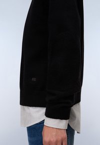 Mustan neuleen paita, jossa on ribbaudet hihansuissa ja teksturoidussa vartalossa, päällä valkoinen raitapaita. Suunnittelu on yksinkertainen ja moderni.