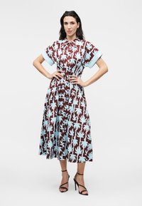 Γυναίκα στέκεται με τα χέρια στους γοφούς φορώντας ένα καφέ μίντι φόρεμα με ανοιχτό μπλε σχέδιο φύλλων φοίνικα και μαύρα σανδάλια με ψηλά τακούνια.