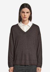 Brauner, übergroßer V-Ausschnitt-Pullover mit gerippten Bündchen und Saum, über einem weißen Hemd mit Kragen getragen. Weiche Textur und lässiges Design.