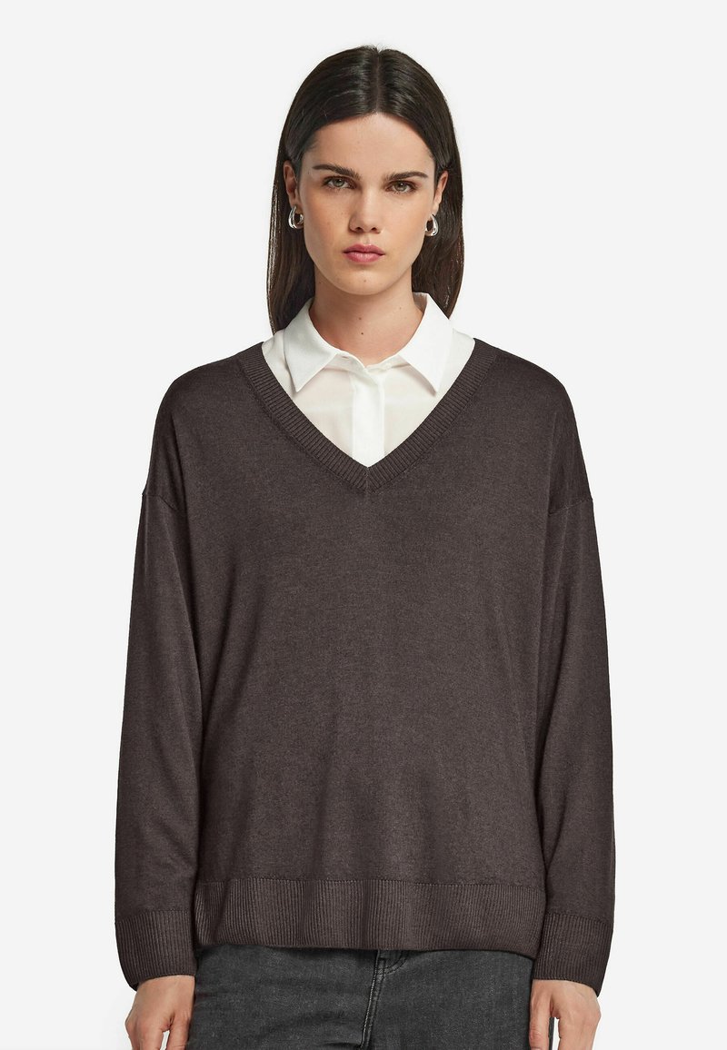 Brauner, übergroßer V-Ausschnitt-Pullover mit gerippten Bündchen und Saum, über einem weißen Hemd mit Kragen getragen. Weiche Textur und lässiges Design.