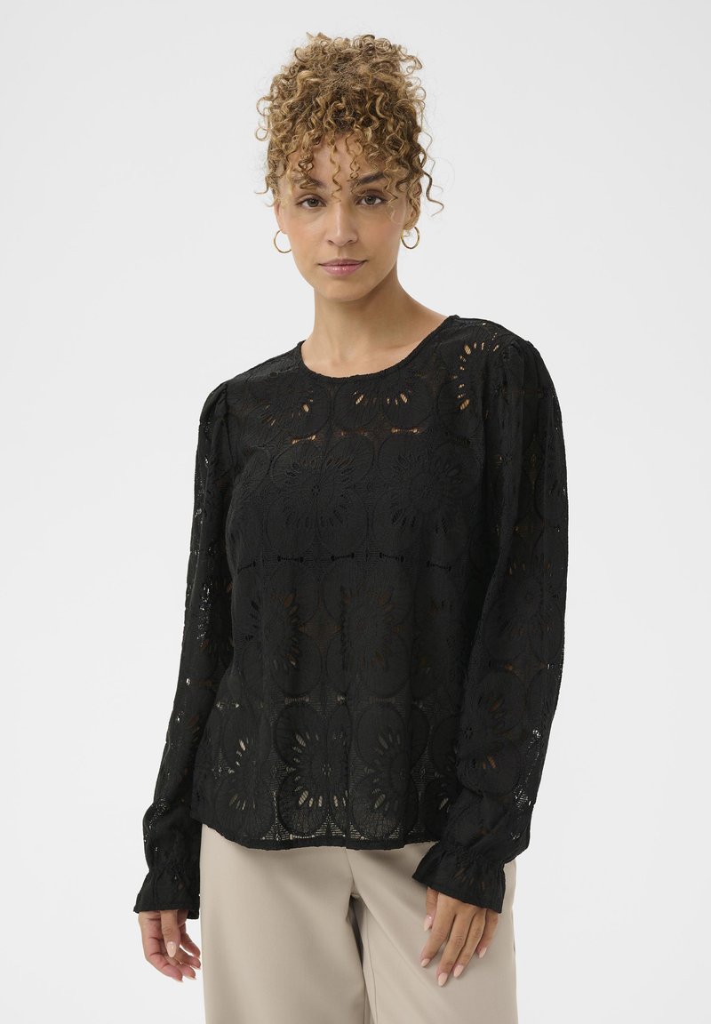 Mujer con cabello rizado, vestida con una blusa negra de encaje floral de manga larga y pantalones beige, de pie contra un fondo claro y liso.