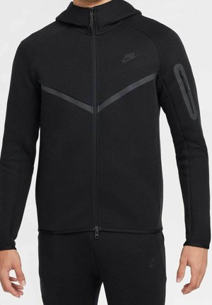 Svart zip-up hoodie med huva, strukturerad tyg, kontrasterande mörka diagonala ränder och en sidoficka med dragkedja på armen.